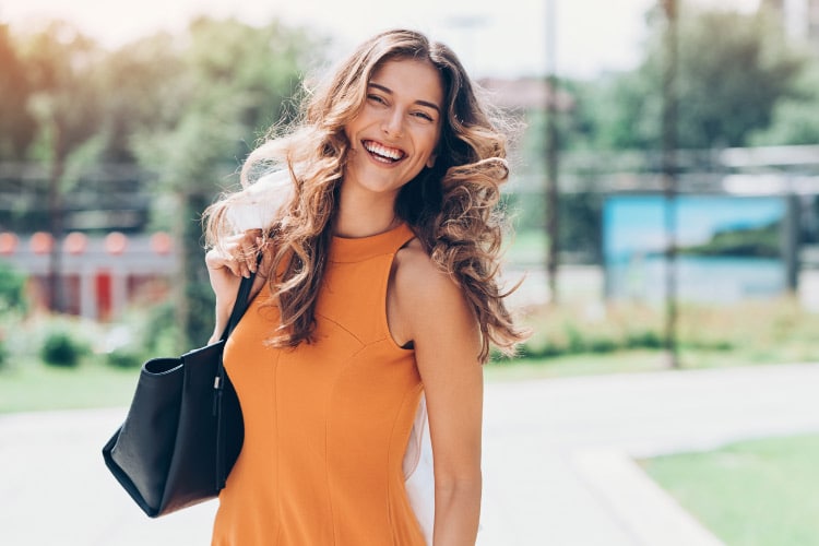 Brunette woman with large curls in an orange dress holds a black purse and smiles outside
